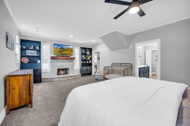 bedroom with ensuite bathroom, a stone fireplace, dark colored carpet, ornamental molding, and ceiling fan