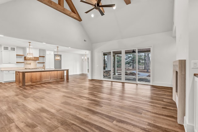 unfurnished living room with high vaulted ceiling, beamed ceiling, light wood-type flooring, a chandelier, and a ceiling fan