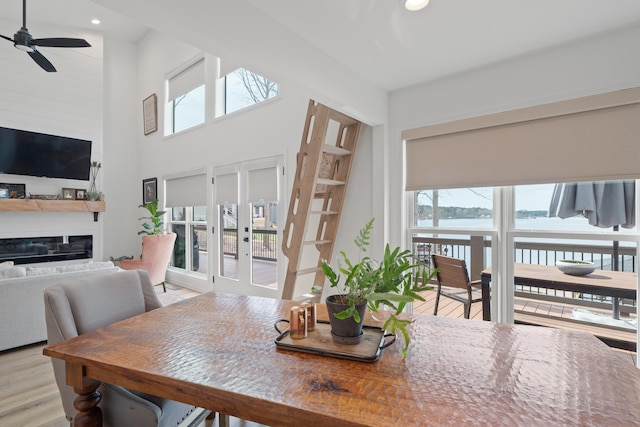 dining space featuring a glass covered fireplace, a towering ceiling, ceiling fan, a water view, and light wood-type flooring