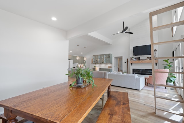 dining space with a ceiling fan, high vaulted ceiling, light wood-style floors, a glass covered fireplace, and beamed ceiling