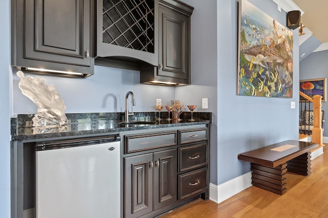 bar featuring sink, stainless steel refrigerator, dark brown cabinetry, light hardwood / wood-style floors, and dark stone counters