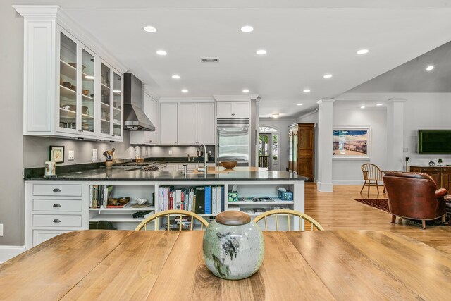 kitchen with wall chimney range hood, stainless steel built in refrigerator, dark countertops, light wood finished floors, and white cabinets