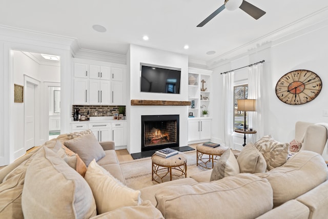 living room featuring crown molding, built in features, a lit fireplace, light wood-style floors, and a ceiling fan