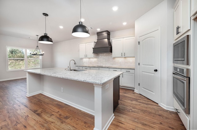 kitchen with premium range hood, appliances with stainless steel finishes, sink, white cabinets, and a kitchen island with sink