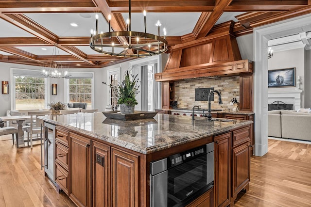 kitchen featuring coffered ceiling, sink, an island with sink, a notable chandelier, and dark stone counters