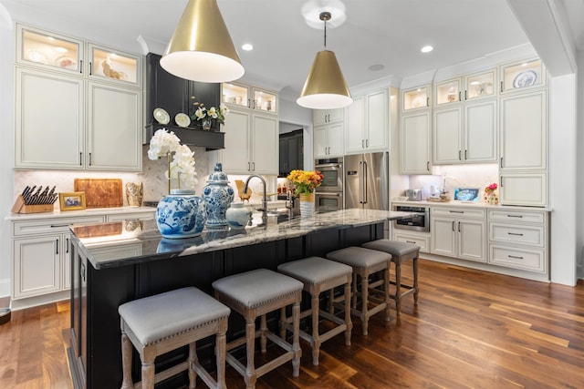 kitchen featuring dark wood-type flooring, a kitchen bar, light stone counters, appliances with stainless steel finishes, and an island with sink