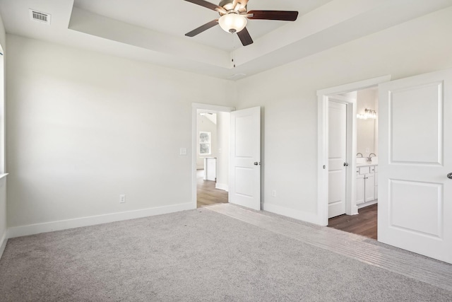 unfurnished bedroom with ceiling fan, ensuite bath, a raised ceiling, and dark colored carpet