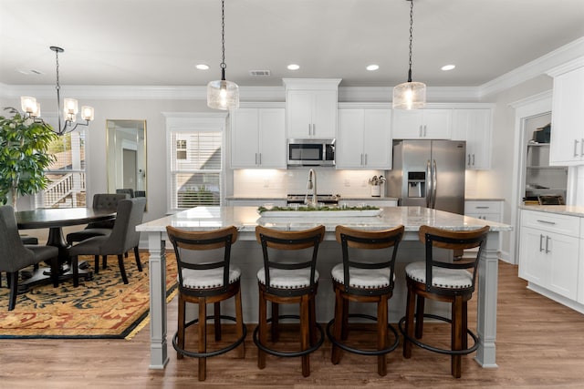 kitchen featuring an island with sink, white cabinets, hanging light fixtures, light stone counters, and stainless steel appliances