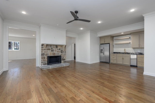 unfurnished living room featuring crown molding, a stone fireplace, wood finished floors, a ceiling fan, and a sink