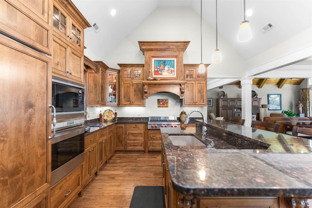 kitchen with a sink, built in appliances, decorative columns, wood finished floors, and brown cabinets