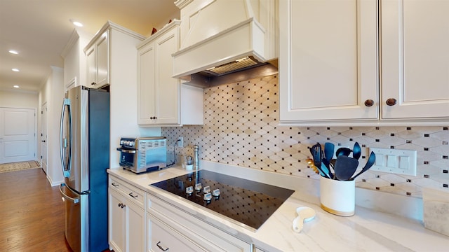 kitchen with stainless steel refrigerator, tasteful backsplash, light stone countertops, custom range hood, and black electric cooktop