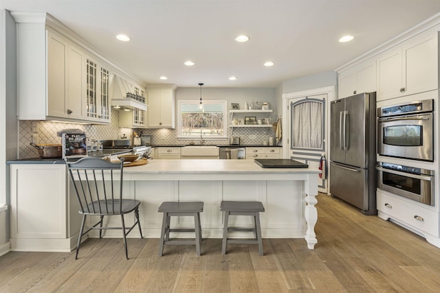 kitchen featuring stainless steel appliances, a peninsula, light wood-style floors, backsplash, and glass insert cabinets