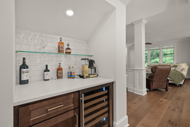 bar with dark wood-type flooring, dark brown cabinets, wine cooler, decorative backsplash, and ornate columns