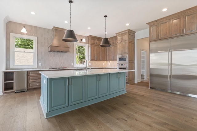 kitchen with decorative light fixtures, a center island with sink, appliances with stainless steel finishes, custom range hood, and light stone countertops