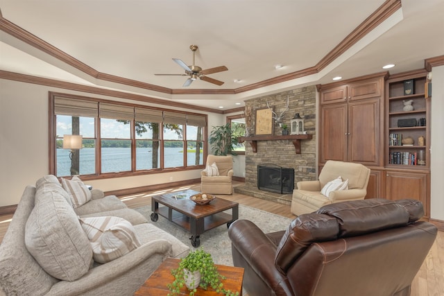 living room featuring a stone fireplace, ceiling fan, a raised ceiling, a water view, and light hardwood / wood-style flooring