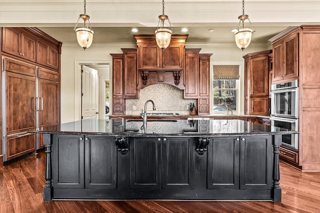 kitchen with dark stone countertops, dark wood-style floors, paneled built in fridge, a large island with sink, and double oven