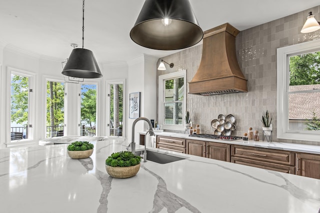 kitchen with a sink, brown cabinetry, premium range hood, light stone counters, and stainless steel gas cooktop