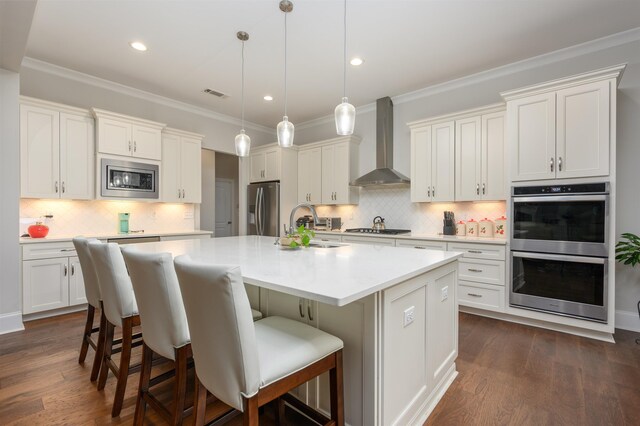 kitchen with sink, appliances with stainless steel finishes, pendant lighting, a kitchen island with sink, and wall chimney range hood