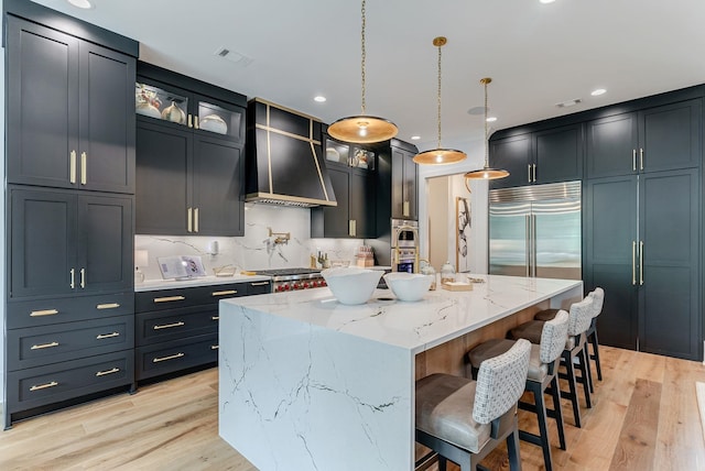 kitchen with decorative light fixtures, a large island with sink, appliances with stainless steel finishes, light stone countertops, and wall chimney range hood