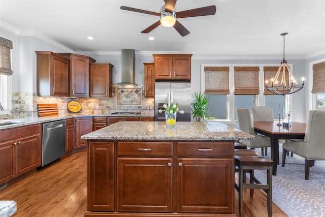 kitchen with tasteful backsplash, wall chimney range hood, ceiling fan with notable chandelier, appliances with stainless steel finishes, and light wood-style flooring