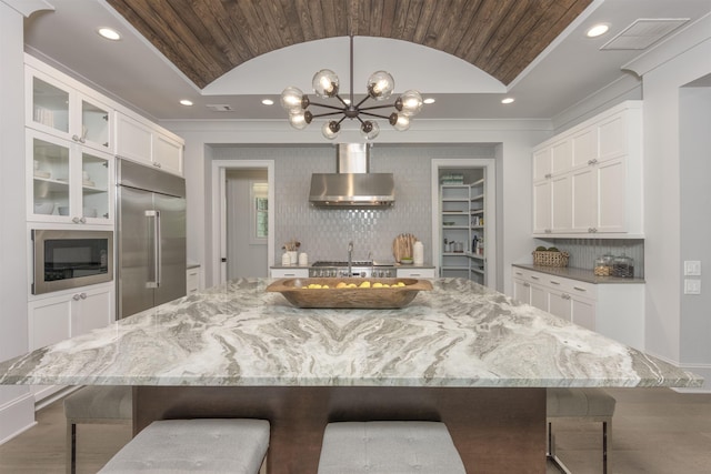 kitchen featuring lofted ceiling, white cabinets, a large island, built in appliances, and wall chimney exhaust hood