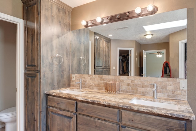 bathroom featuring ceiling fan, toilet, dual bowl vanity, backsplash, and a textured ceiling