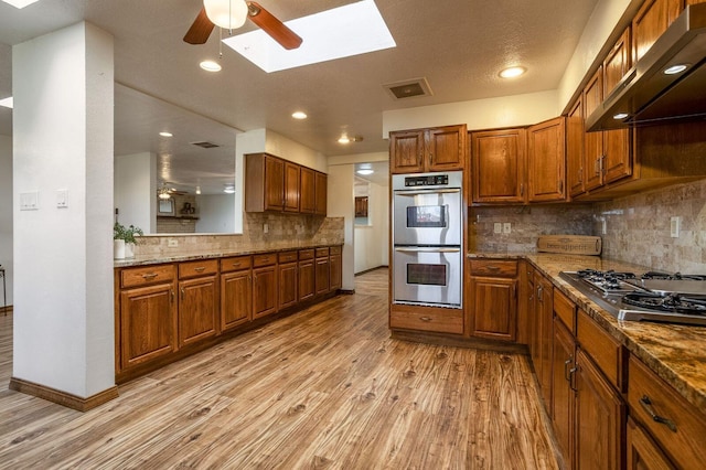 kitchen with appliances with stainless steel finishes, wall chimney range hood, a skylight, light hardwood / wood-style floors, and ceiling fan