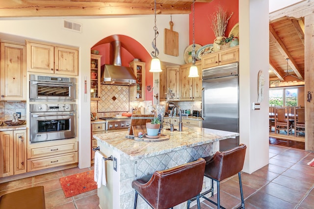 kitchen featuring appliances with stainless steel finishes, hanging light fixtures, light stone counters, a center island with sink, and wall chimney range hood