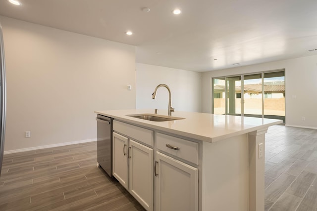 kitchen with wood finish floors, light countertops, stainless steel dishwasher, a kitchen island with sink, and a sink