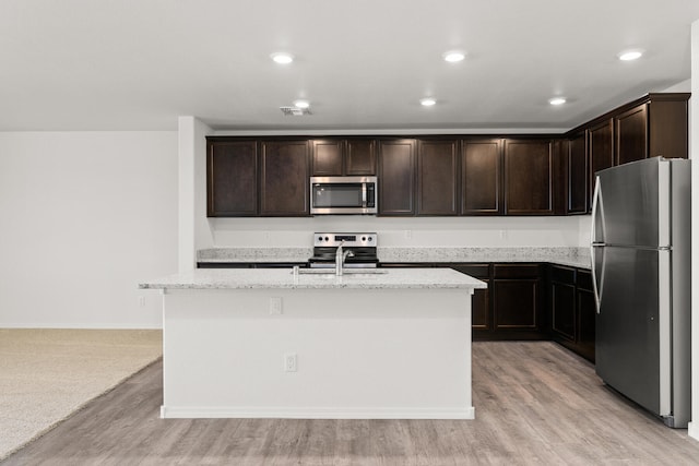 kitchen featuring light stone countertops, a center island with sink, light hardwood / wood-style flooring, appliances with stainless steel finishes, and dark brown cabinetry