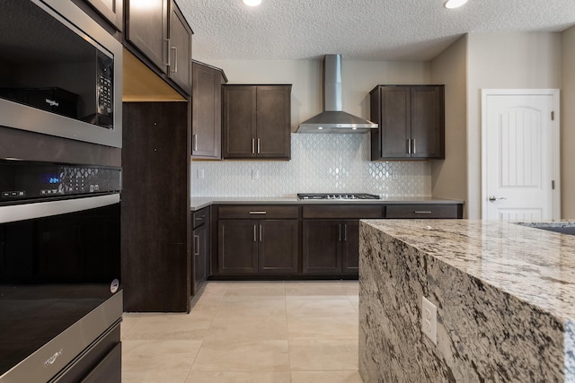 kitchen with wall chimney exhaust hood, dark brown cabinetry, light stone countertops, and stainless steel appliances