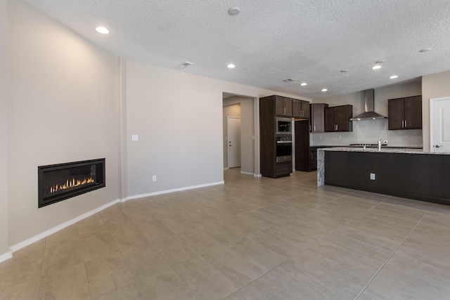 kitchen featuring dark brown cabinetry, wall chimney range hood, light stone counters, backsplash, and a textured ceiling