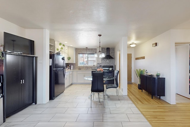 kitchen featuring black fridge, pendant lighting, white cabinets, wall chimney range hood, and backsplash