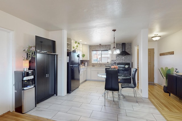 kitchen featuring wall chimney exhaust hood, decorative light fixtures, tasteful backsplash, black refrigerator, and white cabinets