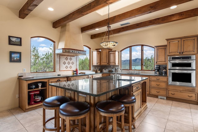 kitchen with sink, stainless steel double oven, dark stone countertops, an island with sink, and custom range hood