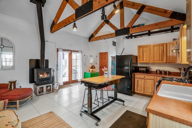 kitchen with light tile patterned flooring, wall chimney range hood, black refrigerator, a wood stove, and high vaulted ceiling