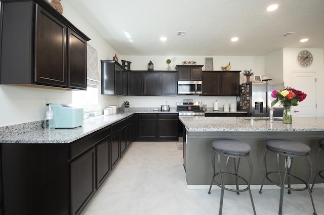 kitchen with a center island with sink, a breakfast bar, dark brown cabinetry, light stone counters, and appliances with stainless steel finishes