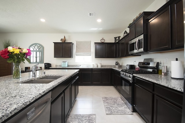 kitchen featuring appliances with stainless steel finishes, dark brown cabinetry, light stone countertops, light tile patterned flooring, and sink