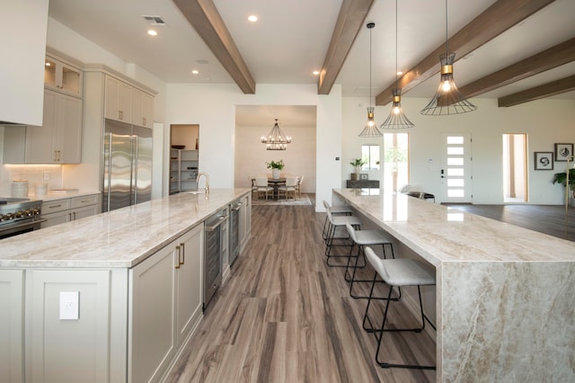 kitchen with a large island with sink, beam ceiling, hanging light fixtures, stainless steel appliances, and hardwood / wood-style floors