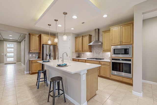 kitchen with a center island with sink, a kitchen breakfast bar, stainless steel appliances, light countertops, and wall chimney range hood