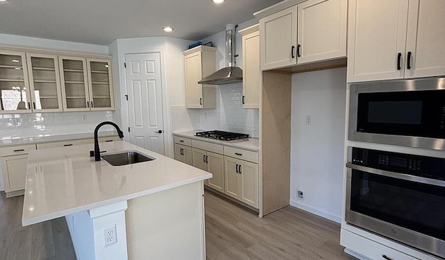 kitchen with a sink, wall chimney range hood, appliances with stainless steel finishes, recessed lighting, and light wood finished floors