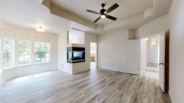 unfurnished living room featuring a tray ceiling, a multi sided fireplace, ceiling fan, and light hardwood / wood-style floors