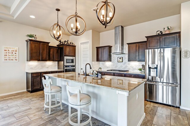 kitchen with wall chimney exhaust hood, a notable chandelier, a kitchen island with sink, stainless steel appliances, and light stone counters