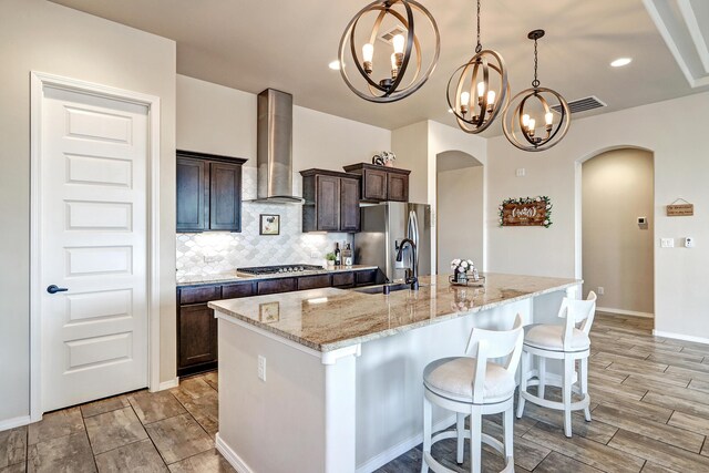 kitchen with decorative light fixtures, a chandelier, sink, wall chimney exhaust hood, and an island with sink