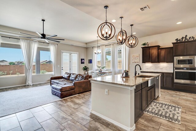 kitchen featuring ceiling fan with notable chandelier, appliances with stainless steel finishes, light stone countertops, sink, and a center island with sink