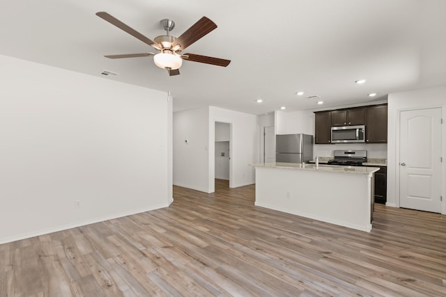 kitchen featuring a kitchen island with sink, stainless steel appliances, ceiling fan, light stone counters, and light wood-type flooring