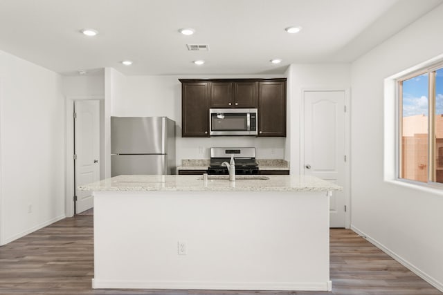 kitchen with appliances with stainless steel finishes, light stone counters, light hardwood / wood-style floors, dark brown cabinets, and a center island with sink