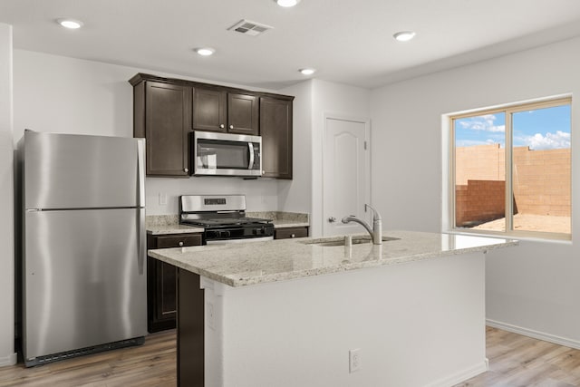 kitchen with light stone countertops, a center island with sink, appliances with stainless steel finishes, sink, and light wood-type flooring