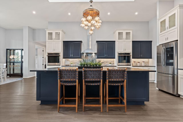 kitchen with a spacious island, a notable chandelier, appliances with stainless steel finishes, dark wood-type flooring, and wall chimney range hood