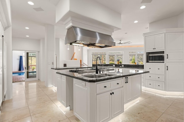 kitchen with light tile patterned floors, stainless steel microwave, a kitchen island with sink, a sink, and wall chimney exhaust hood